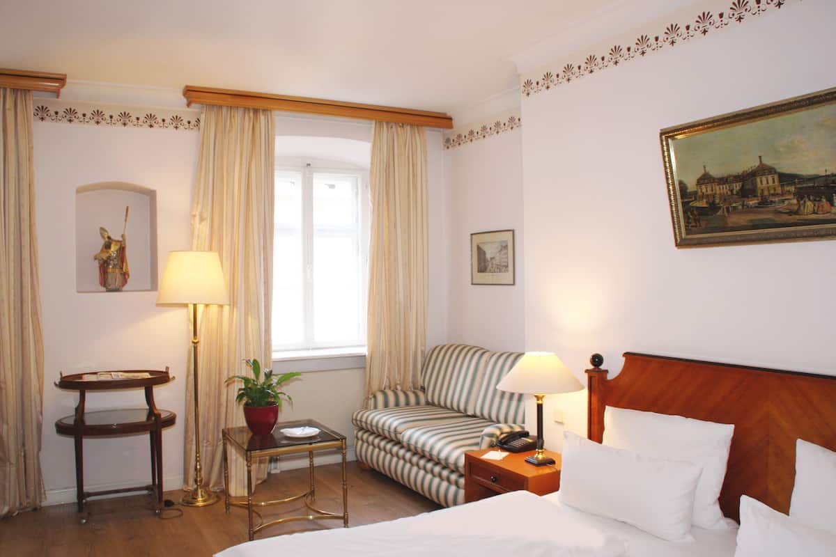 A hotel room with a bed, a striped sofa, a small table with a lamp, a potted plant, and a painting on the wall.
