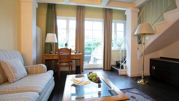 Een woonkamer met een bank, een glazen bijzettafeltje met fruit, een lamp, een bureau en uitzicht op de buitenlucht door grote ramen.