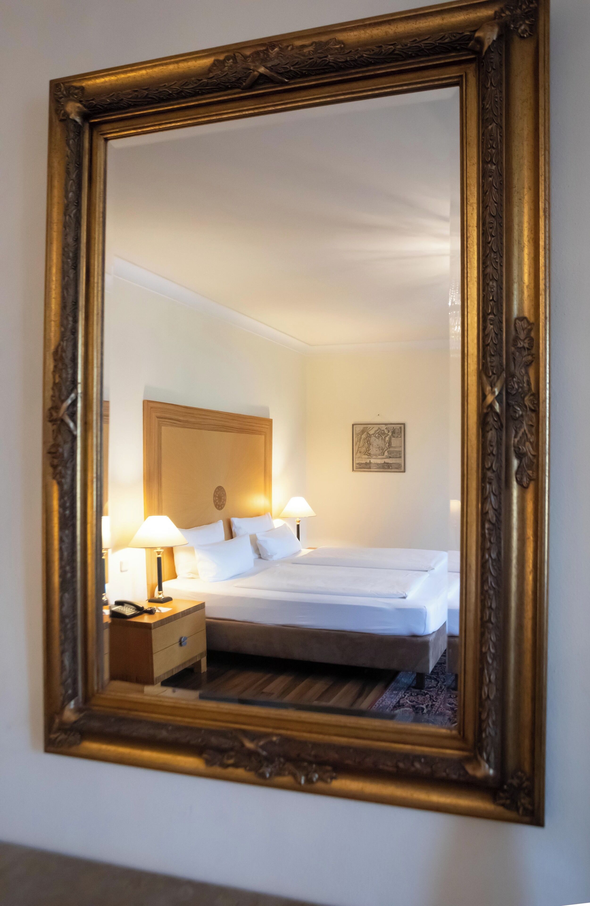 A hotel room with a large ornate mirror, a bed with white linens, two bedside lamps, a wooden nightstand, and a framed picture on the wall.