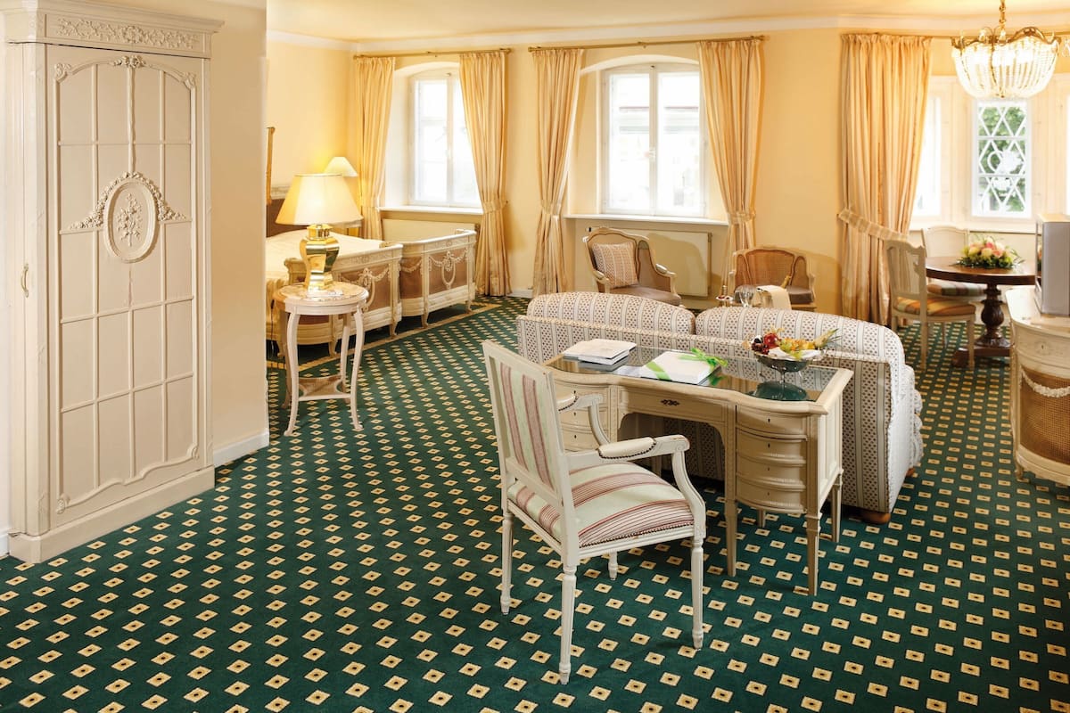 A hotel room with a green patterned carpet, a dining table with chairs, a bed with a canopy, a dresser, and a large armoire.