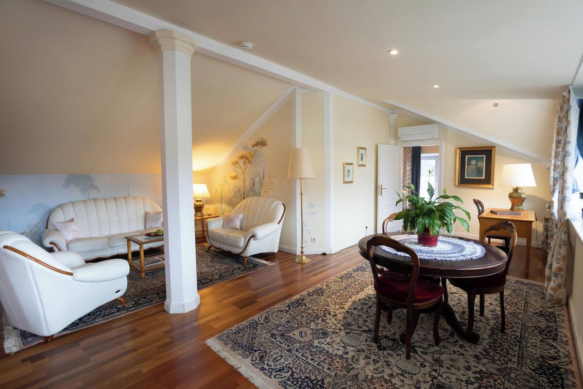 A spacious living room with a white sofa, a white armchair, a dining table with chairs, and a large patterned rug.