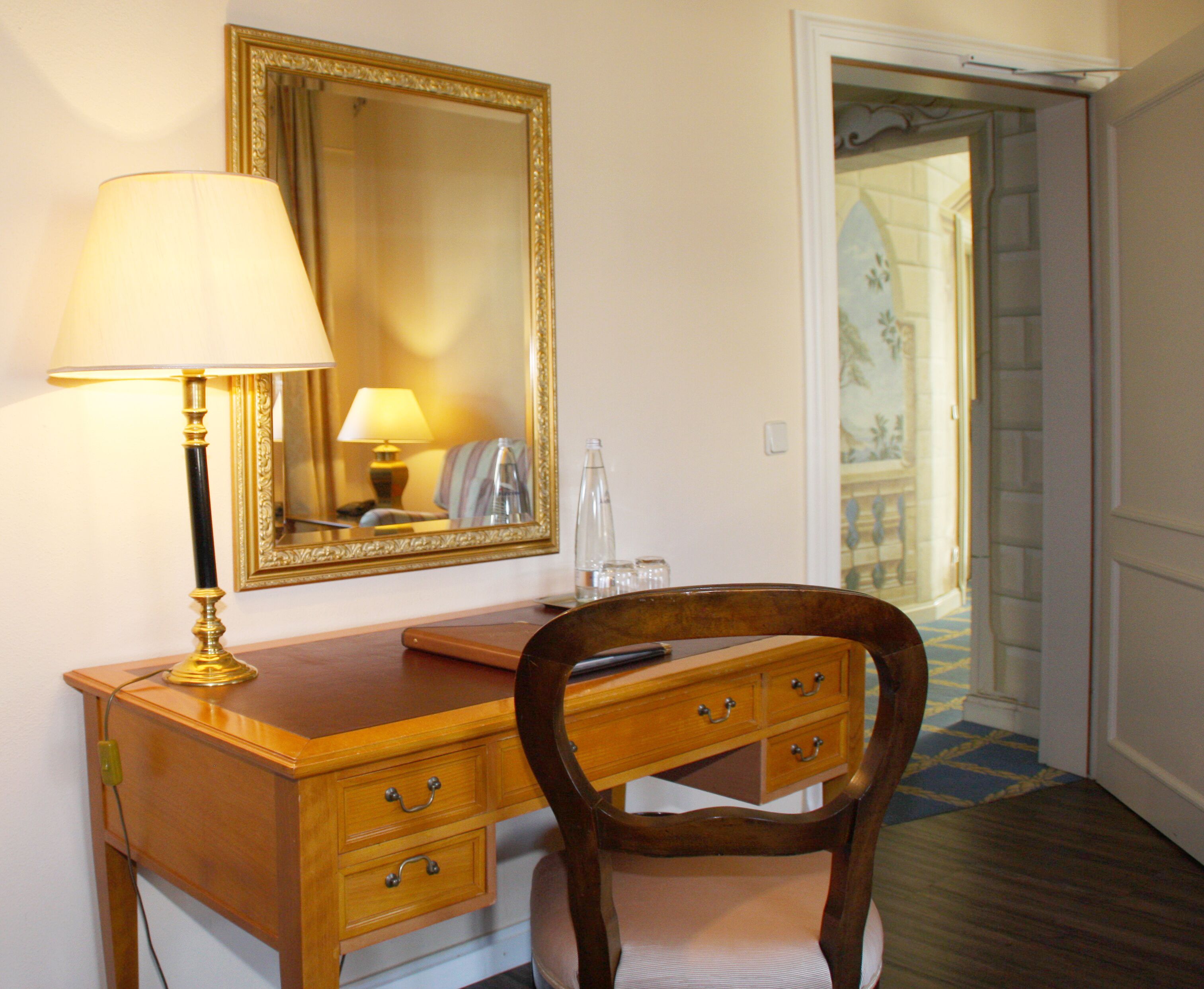A wooden desk with a lamp, a mirror, and a chair in a room with a door leading to a balcony.