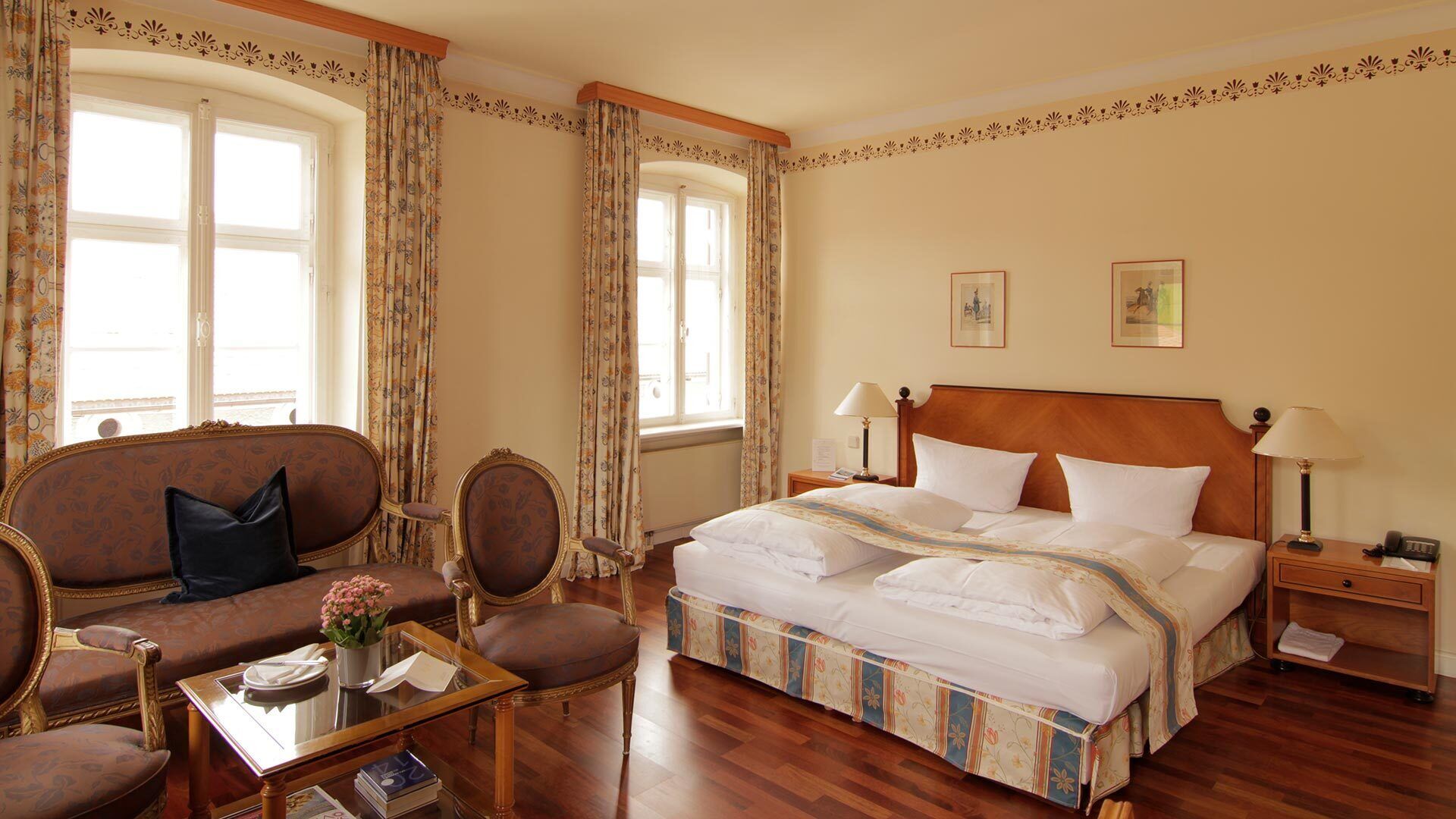 Ein Hotelzimmer mit einem großen Bett, zwei Nachttischen, einem hölzernen Kopfteil, einem Sofa, einem Sessel und einem kleinen Tisch mit Blumen in einer Vase.