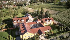Un edificio grande de varios pisos con techo de tejas rojas, rodeado de jardines cuidados y viñedos.