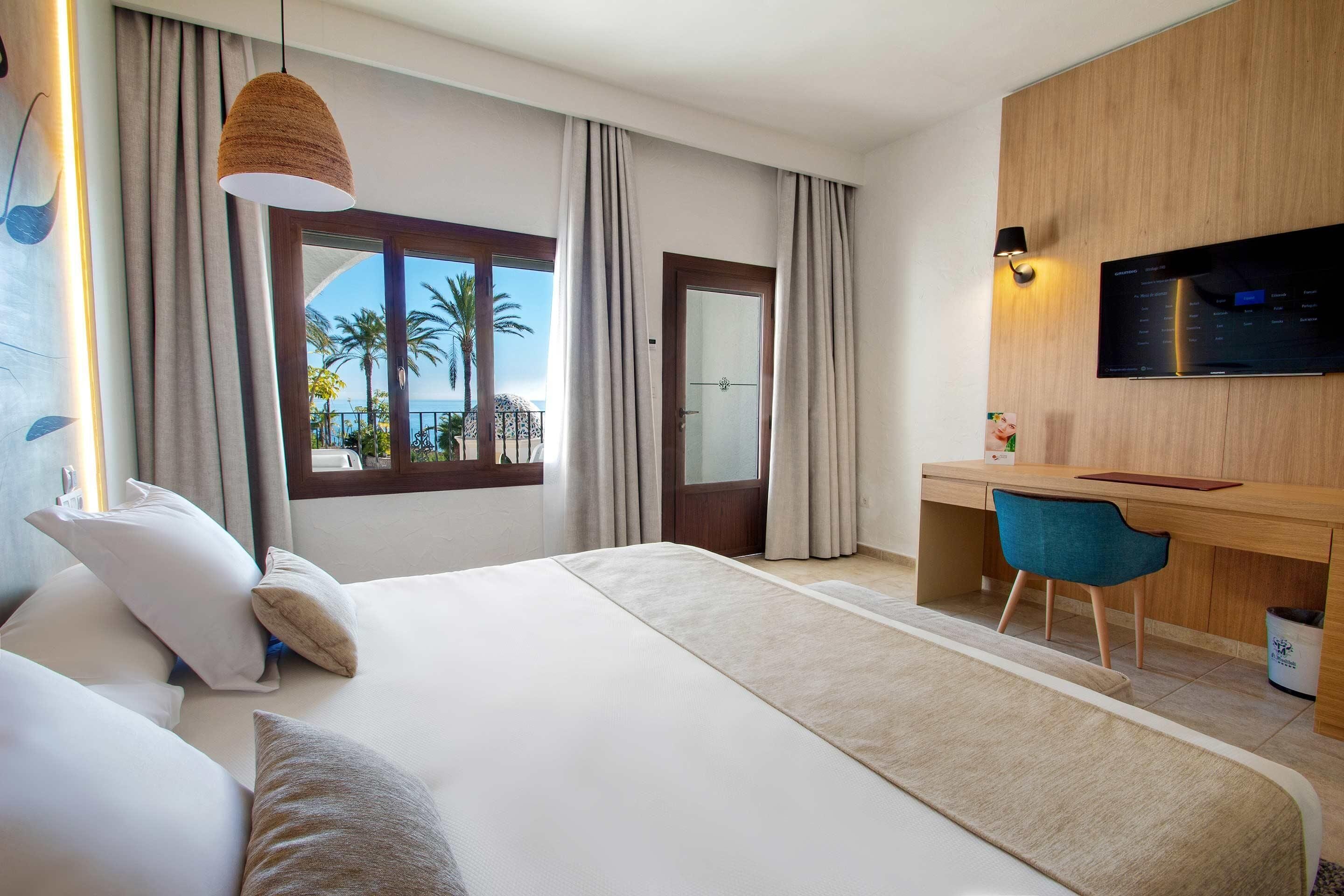A hotel room with a bed, a desk, a chair, a TV, and a view of palm trees and the sea.