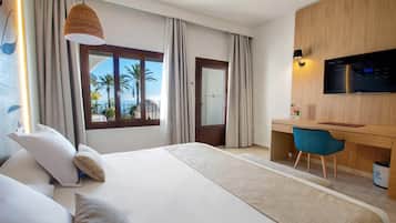 A hotel room with a bed, a desk, a chair, a TV, and a view of palm trees and the sea.