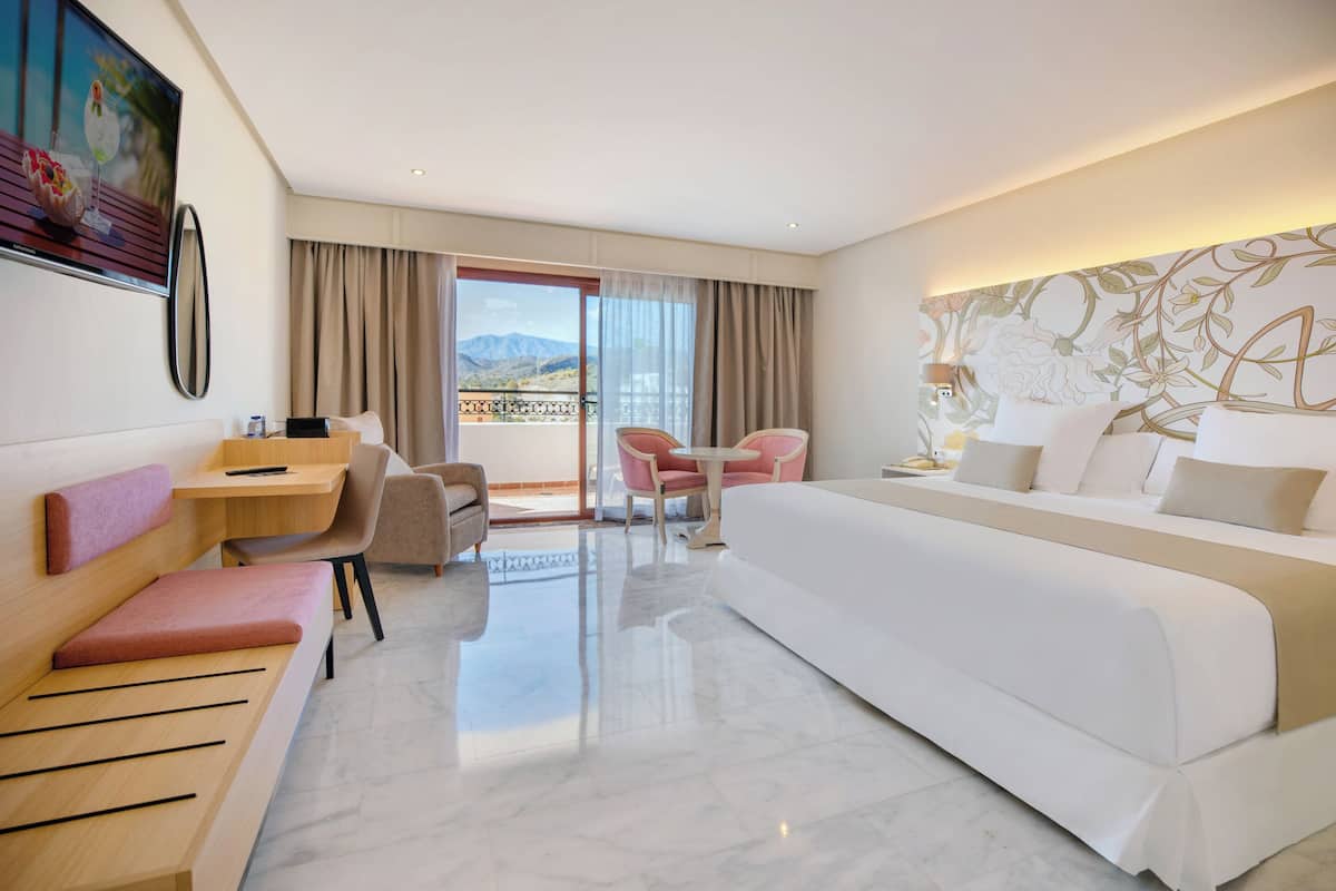 Een hotelkamer met een bed, een bureau, een stoel, een bank en een balkon met uitzicht op de bergen.