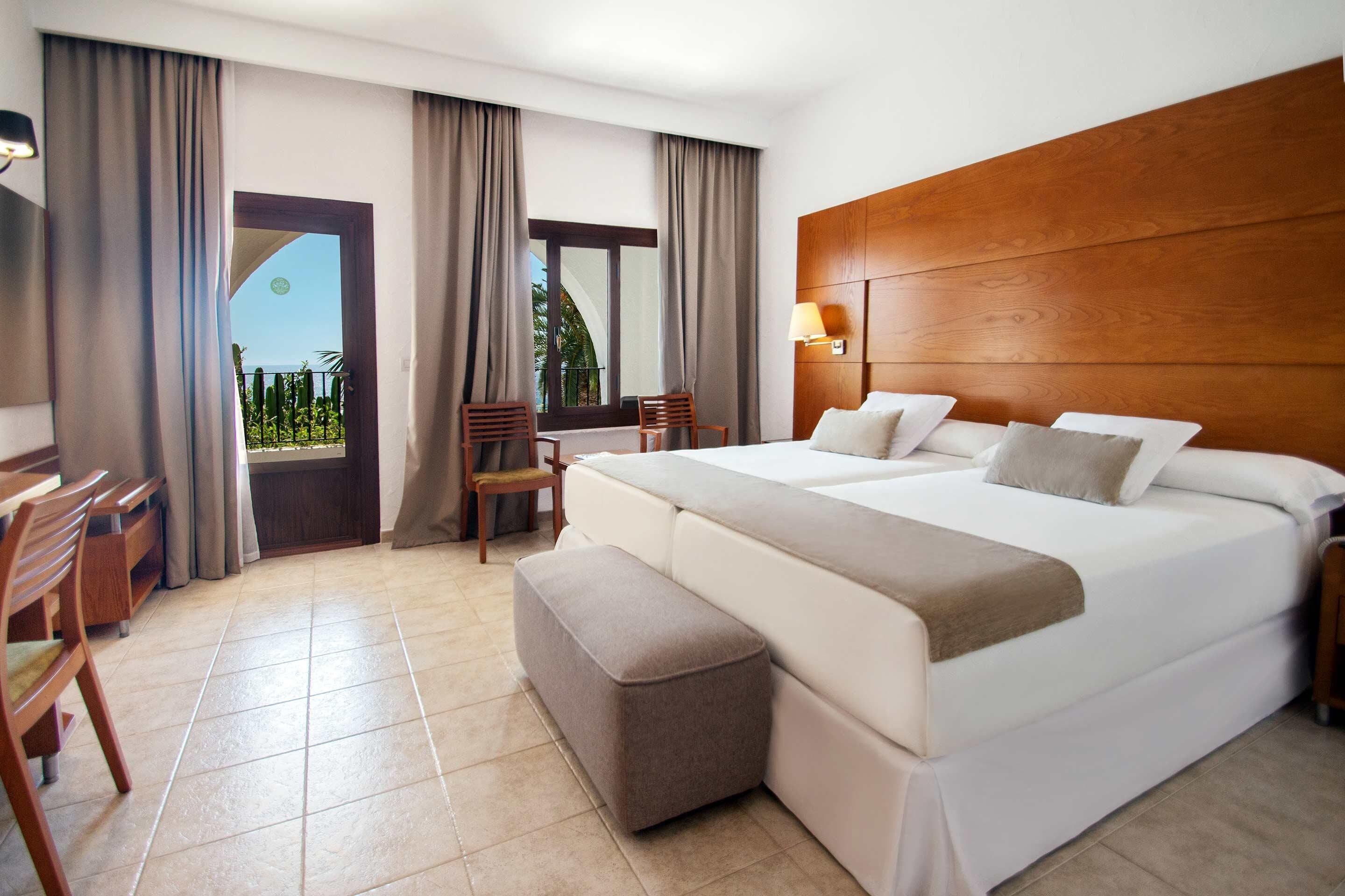 Habitación de hotel con una cama grande, cabecera de madera, un escritorio con sillas, una ventana con cortinas y vistas a la vegetación.