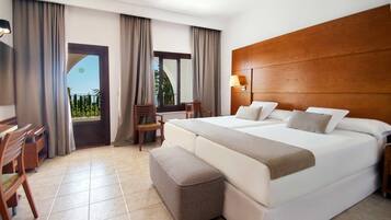 Habitación de hotel con una cama grande, cabecera de madera, un escritorio con sillas, una ventana con cortinas y vistas a la vegetación.