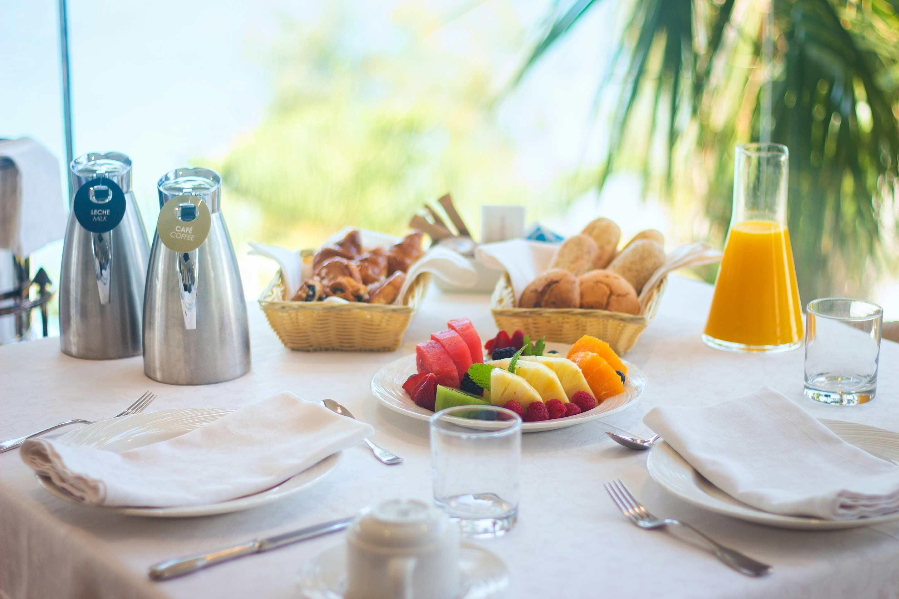 Una mesa con una variedad de frutas, bollería y bebidas para el desayuno.