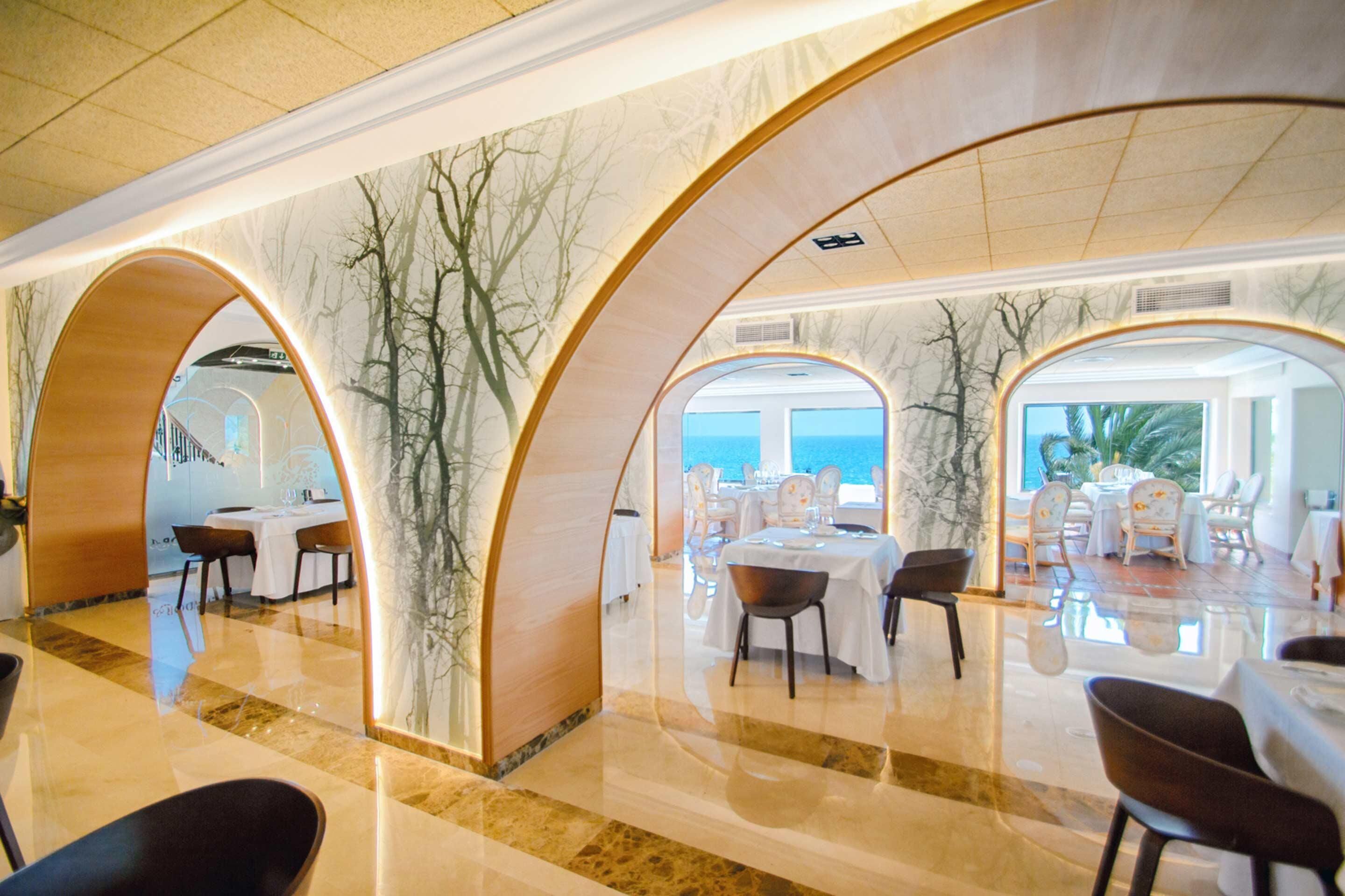 Un ristorante con interni moderni, caratterizzato da pareti e archi in marmo. I tavoli sono apparecchiati per la cena, e l'ambiente è ben illuminato.