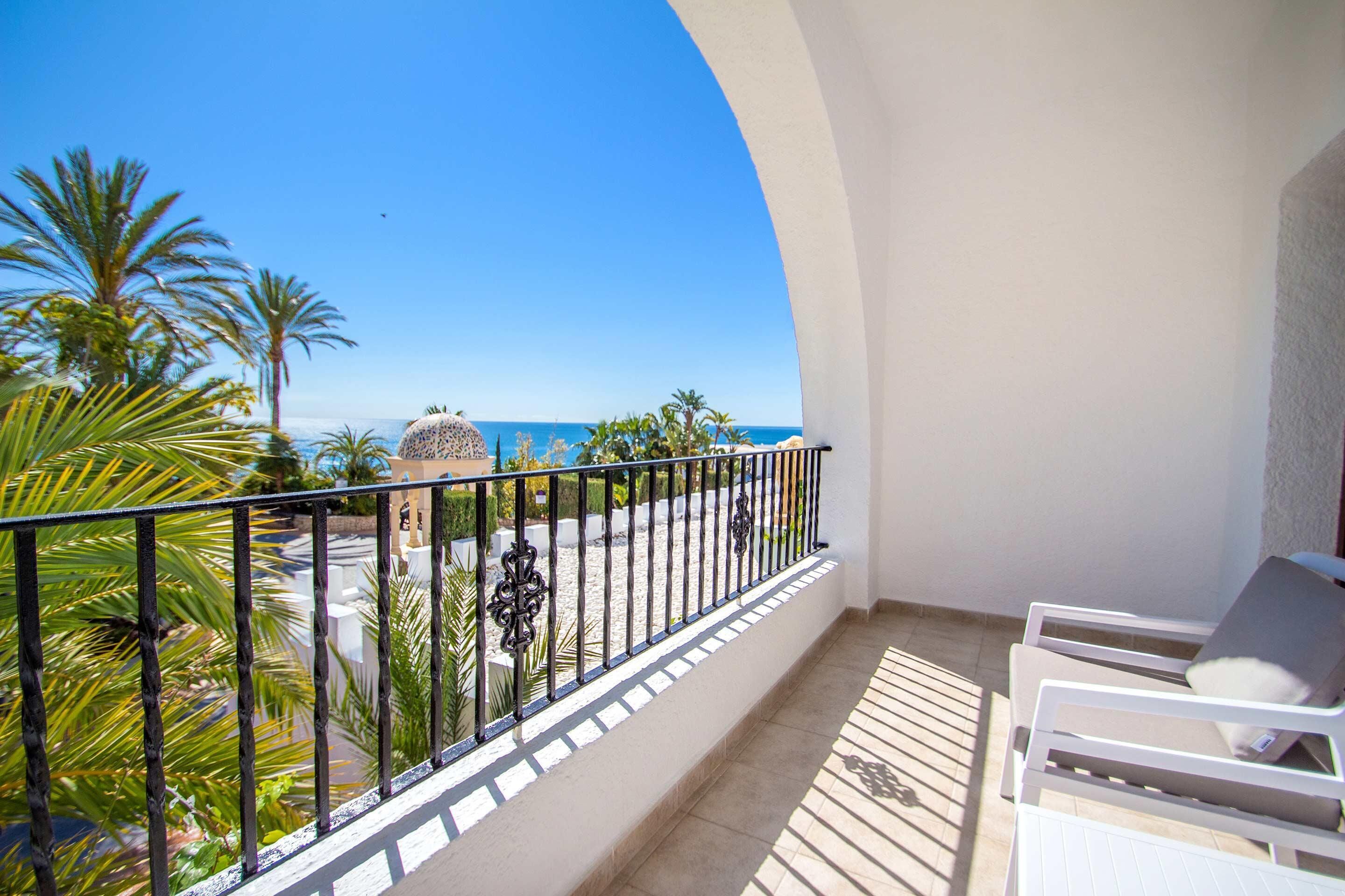 A balcony with a view of the sea, palm trees, and a white building.