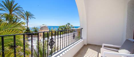 A balcony with a view of the sea, palm trees, and a white building.