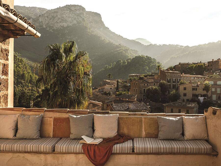 A balcony with a cushioned seating area, overlooking a historic town with traditional buildings and a mountainous backdrop.