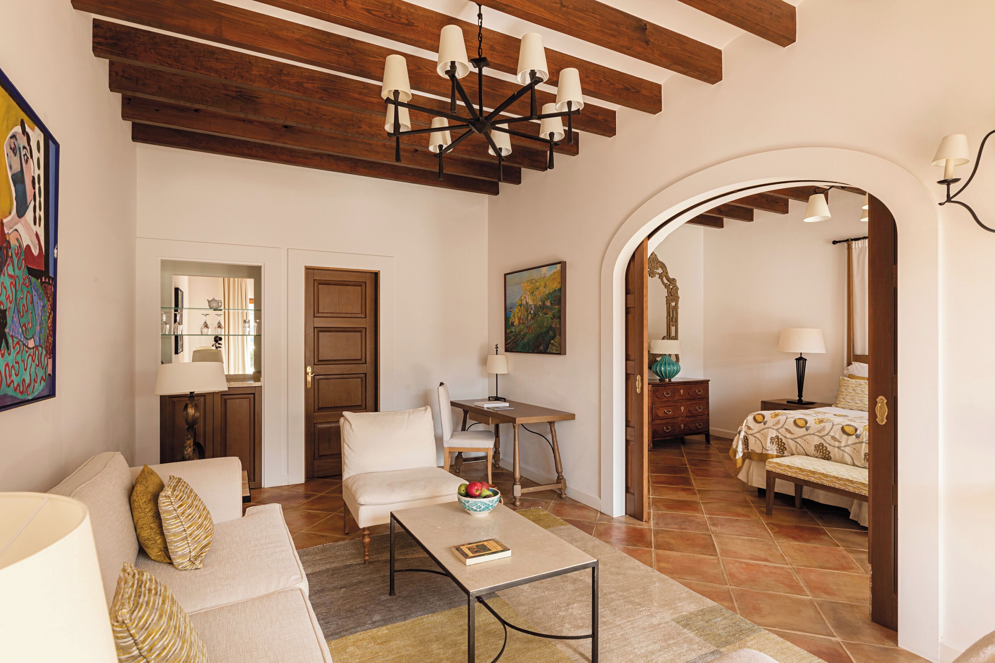 A living room with a sofa, coffee table, and a dining area with a table and chairs. There is a bedroom visible through an archway with a bed and a nightstand.