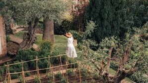 A garden with a person in a white dress, a large tree, and buildings with tiled roofs in the background.