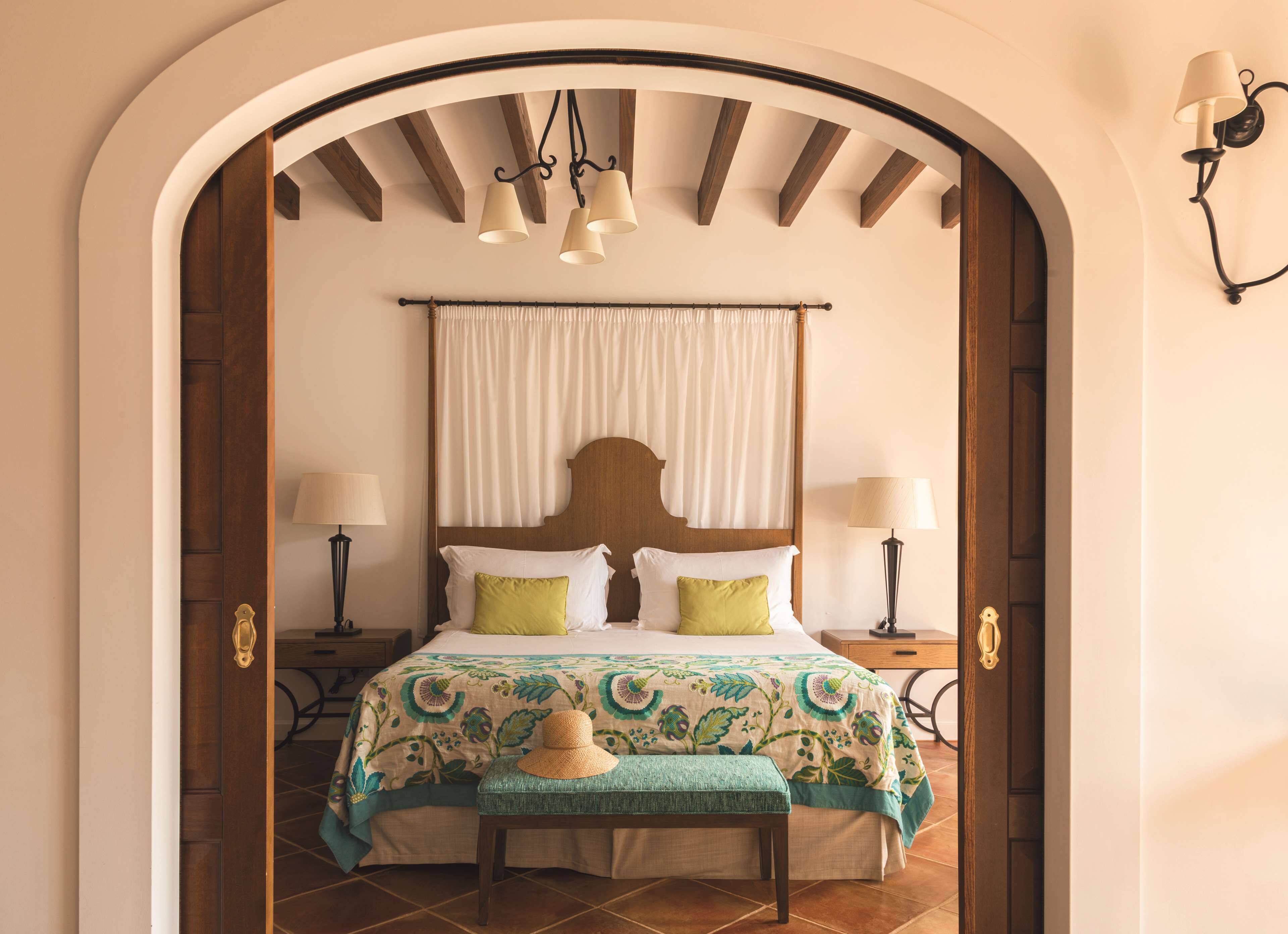 Un dormitorio con cabecera de madera, dos mesitas de noche con lámparas, una cama con colcha estampada, un banco y un arco de entrada.