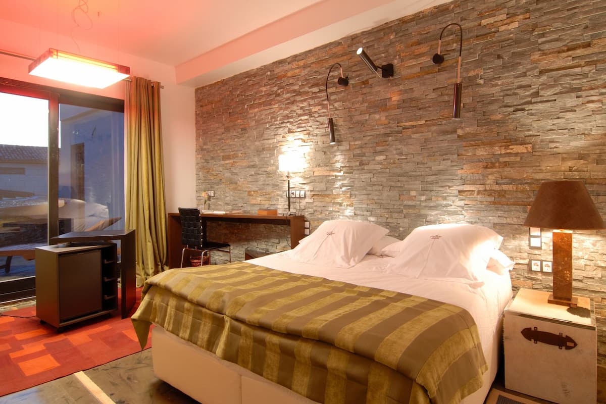 Quarto de hotel moderno com parede de pedra, cama com lençóis brancos, escrivaninha com cadeira e uma janela grande com cortinas.