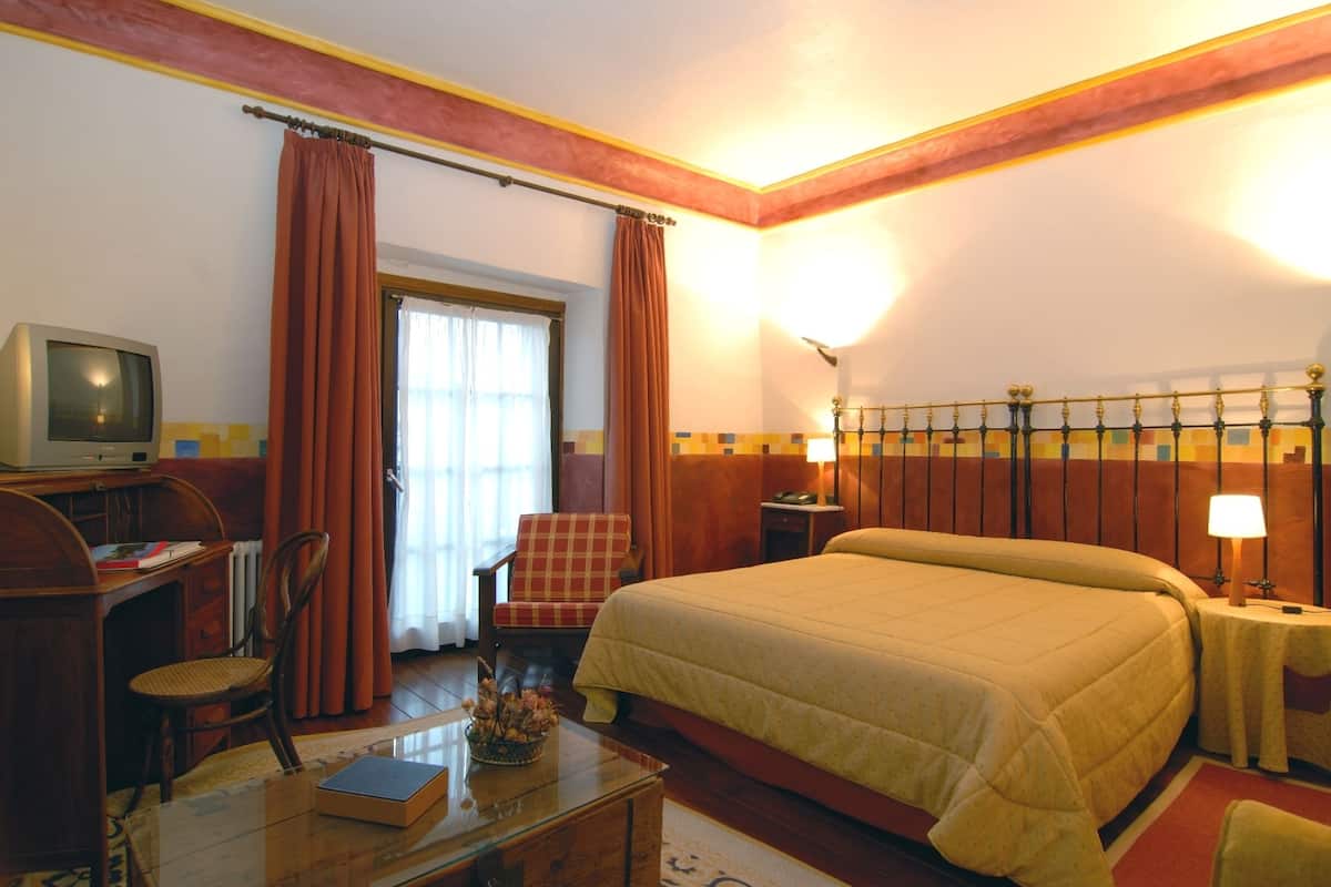 Quarto de hotel com uma cama, uma mesa com televisão, uma cadeira, um abajur e uma janela com cortinas.