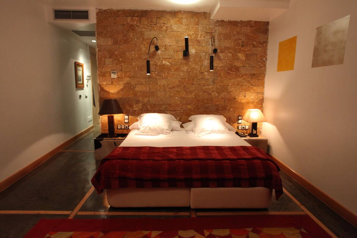 Quarto de hotel com parede de tijolos, cama com roupa de cama branca, duas luminárias de cabeceira e um cobertor xadrez vermelho e branco.
