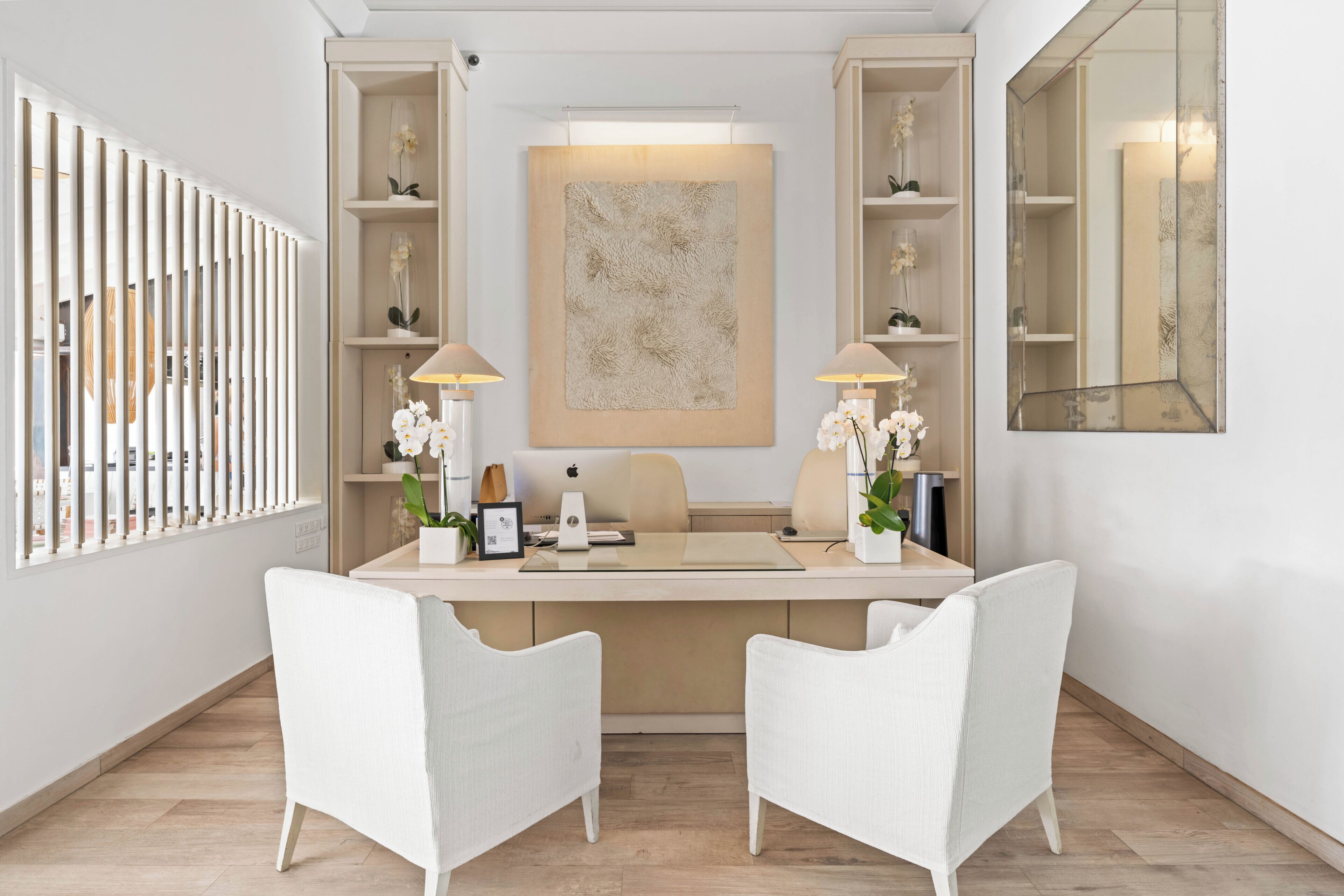 A modern office space with a desk, two chairs, a large mirror, and shelves with decorative items.