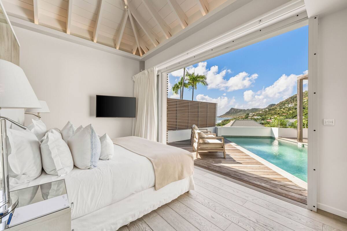 A bedroom with a bed, a TV, a bedside table, and a view of a pool and mountains.