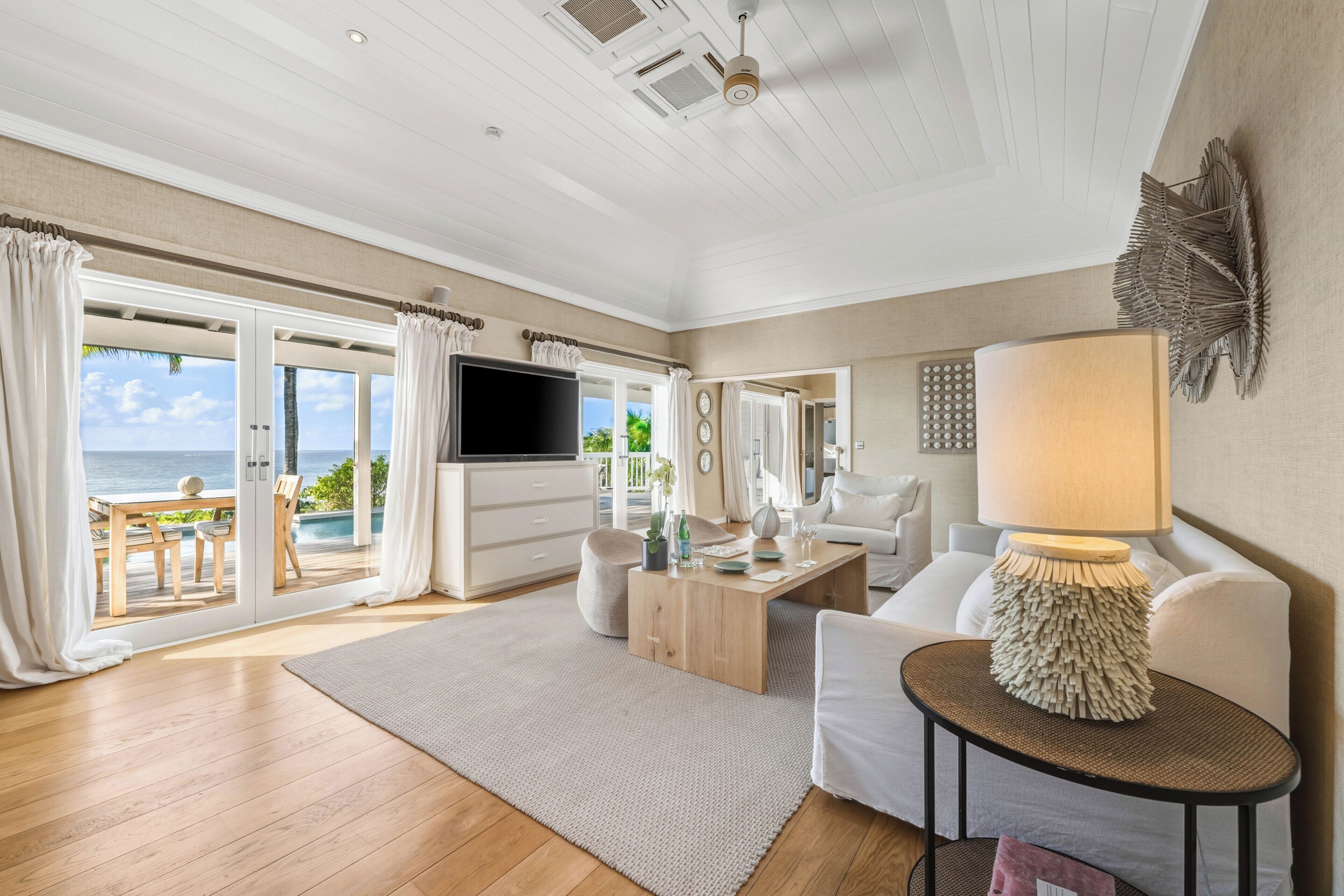 A spacious living room with a wooden floor, a sofa, a coffee table, a TV, and a balcony with a view of the sea.