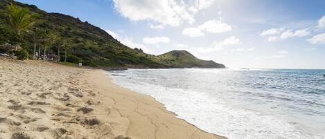Una playa con huellas en la arena, cielo despejado y una montaña al fondo.