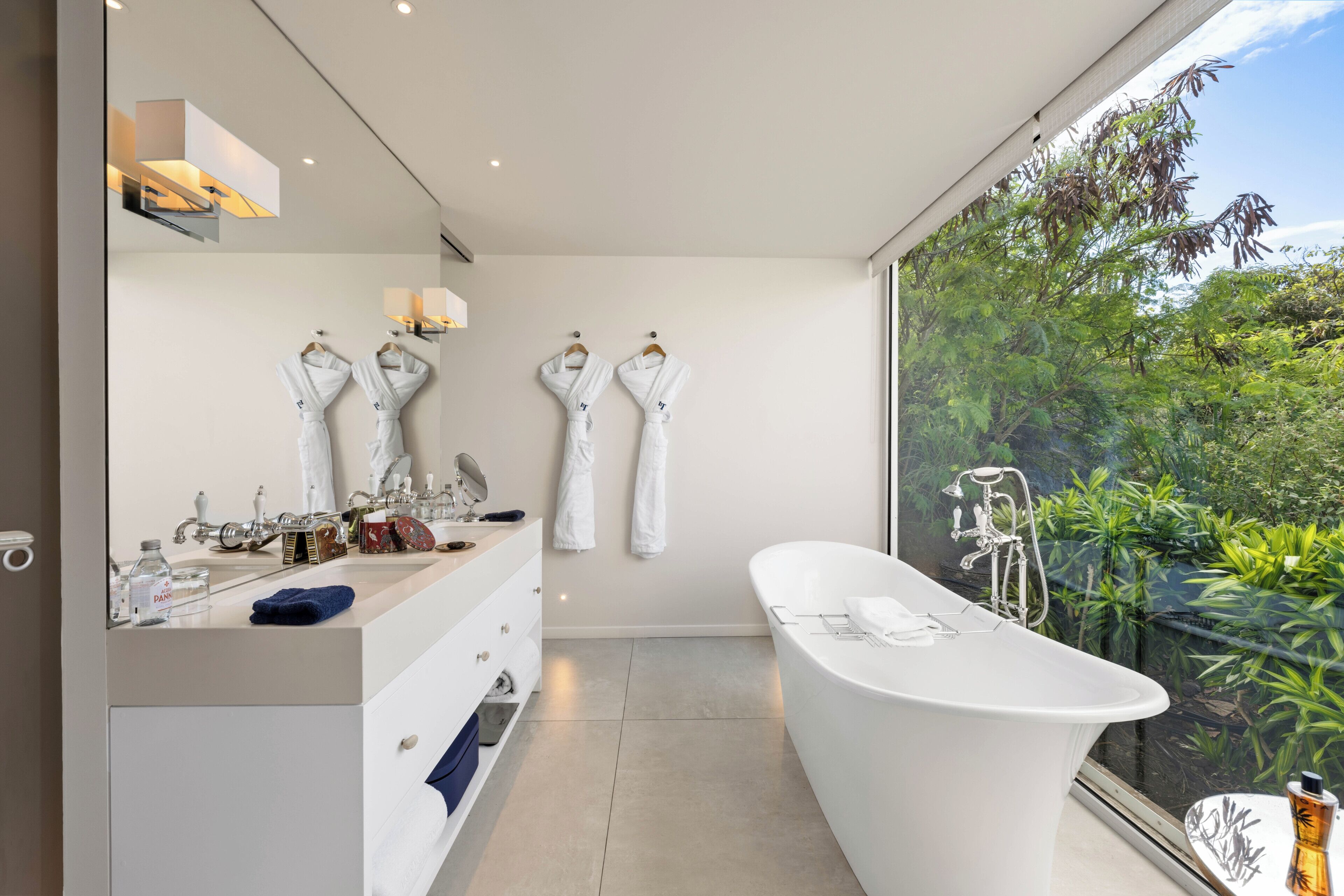 A modern bathroom with a large bathtub, a vanity with a mirror, and a view of greenery outside.