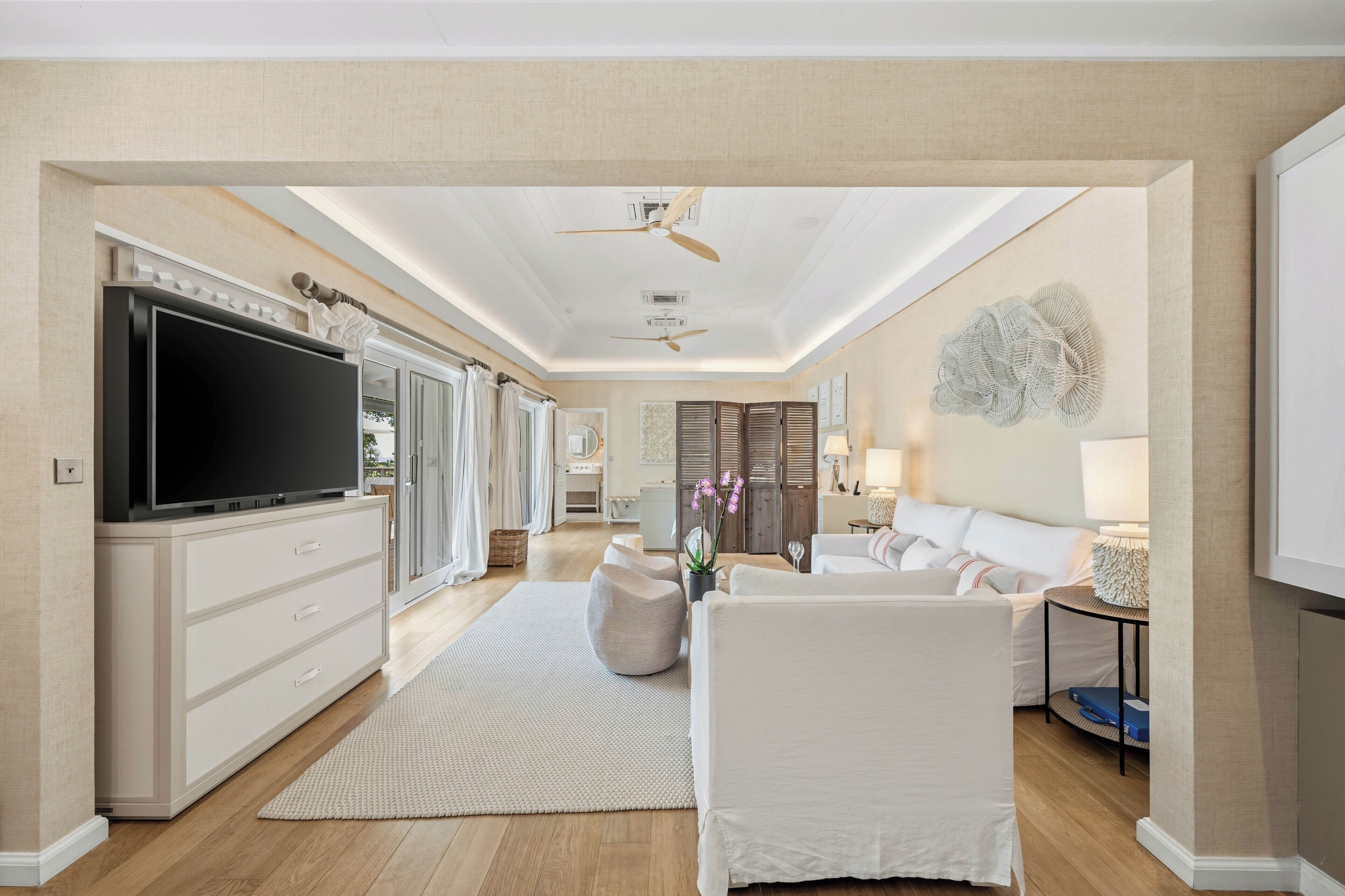 A spacious living area with a flat-screen TV, white furniture, a ceiling fan, and a decorative wall piece.