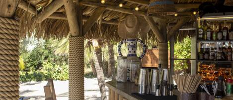 A wooden bar area with striped seating, a thatched roof, and a shelf with bottles.