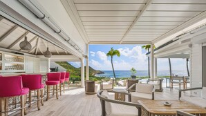 A covered outdoor seating area with a view of the ocean, bar with stools, and a poolside lounge. - Le Toiny Hôtel & Beach Club (St. Barthelemy)