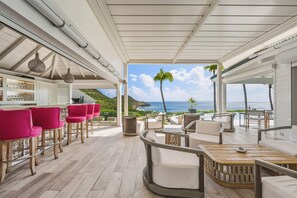 Et overdækket udeområde med havudsigt, bar med barstole og solstole ved poolen.