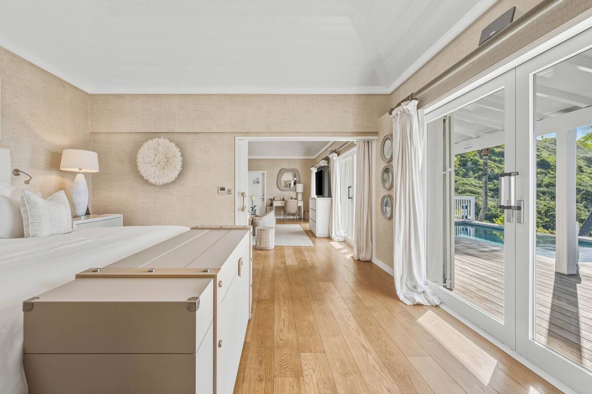 A bedroom with a large bed, a dresser, a nightstand, a lamp, and a view of a balcony with a pool.