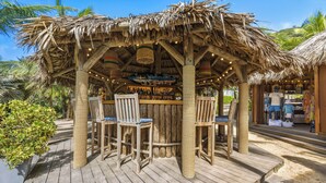 A wooden deck with a thatched roof bar area, surrounded by palm trees and a sandy ground. - Le Toiny Hôtel & Beach Club (St. Barthelemy)