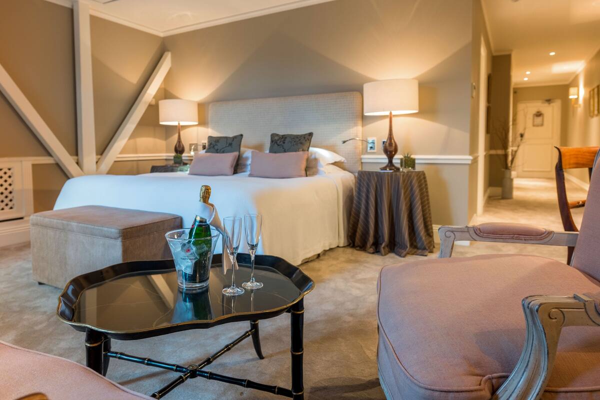 A hotel room with a large bed, a glass-topped table with a bottle and glasses, a chair, and a small side table with a lamp.