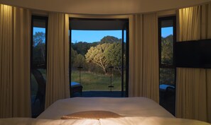 Ett sovrum med en stor säng, utsikt över ett träd genom ett fönster och gardiner på sidorna.