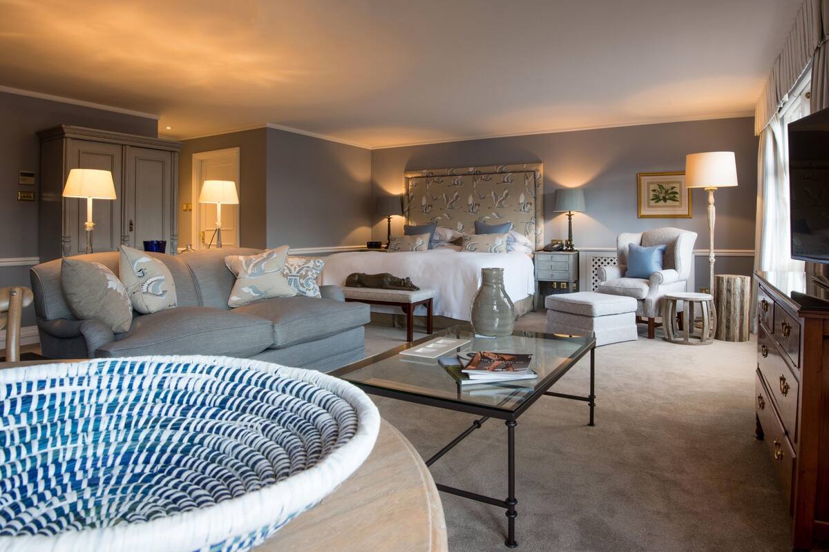 A well-lit living room with a sofa, armchair, and coffee table. A bed with a large headboard is in the background.