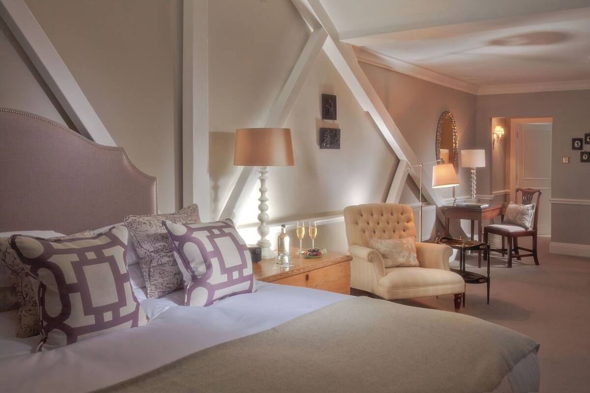 A hotel room with a large bed, a sitting area with a chair, a small table, and a lamp. There is a staircase in the background.