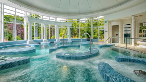 A large indoor swimming pool with multiple levels and a glass ceiling, surrounded by white columns and large windows. - Chewton Glen Hotel & Spa (New Milton)