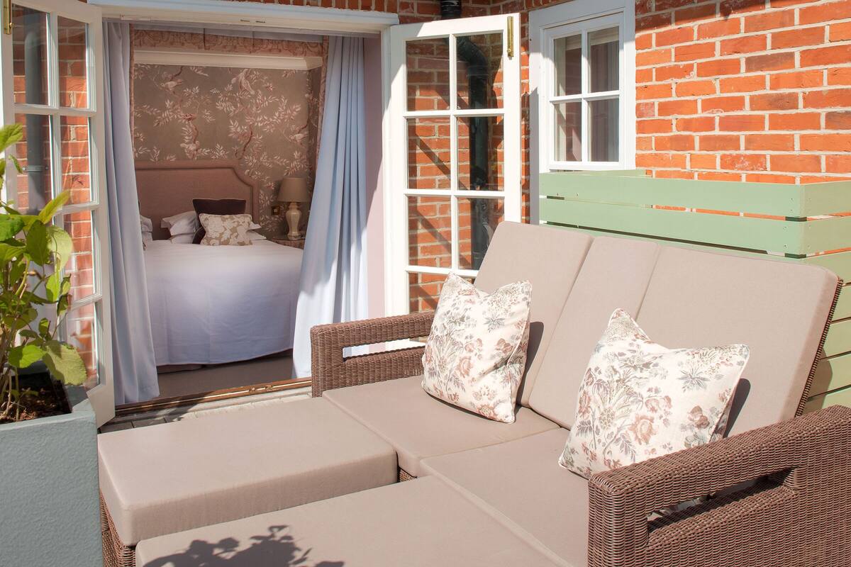 A cozy outdoor seating area with a wicker sofa and cushions, a bed with floral wallpaper, and a brick wall.