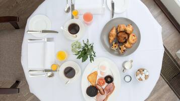 A breakfast setup on a round table with plates of eggs, bacon, tomatoes, and toast, accompanied by coffee, juice, and pastries.
