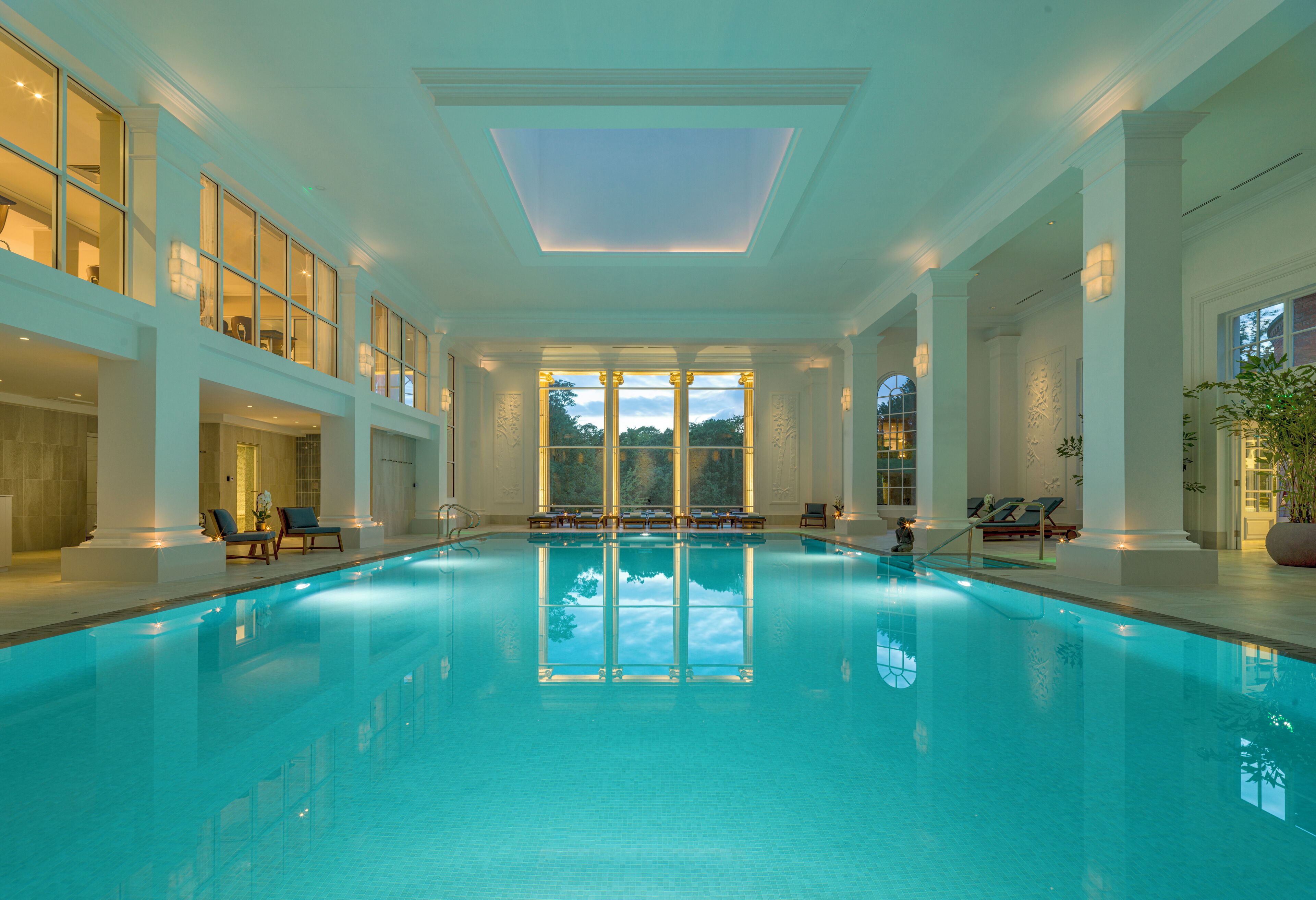 A large indoor swimming pool with clear glass walls, multiple lounge areas, and a skylight.