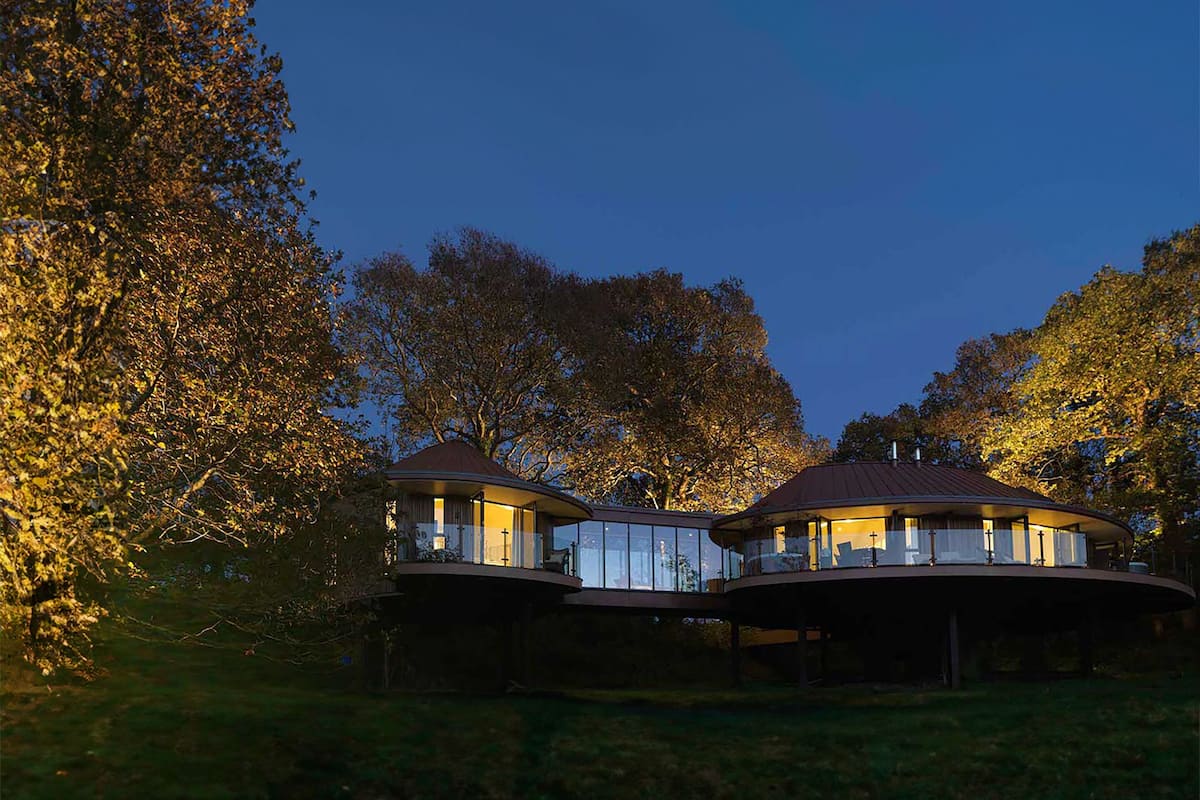 A modern house with two separate sections, each with a balcony and large windows, surrounded by trees.