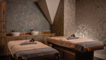 A spa room with two massage tables, a wooden cabinet, a sink, and a tiled wall.
