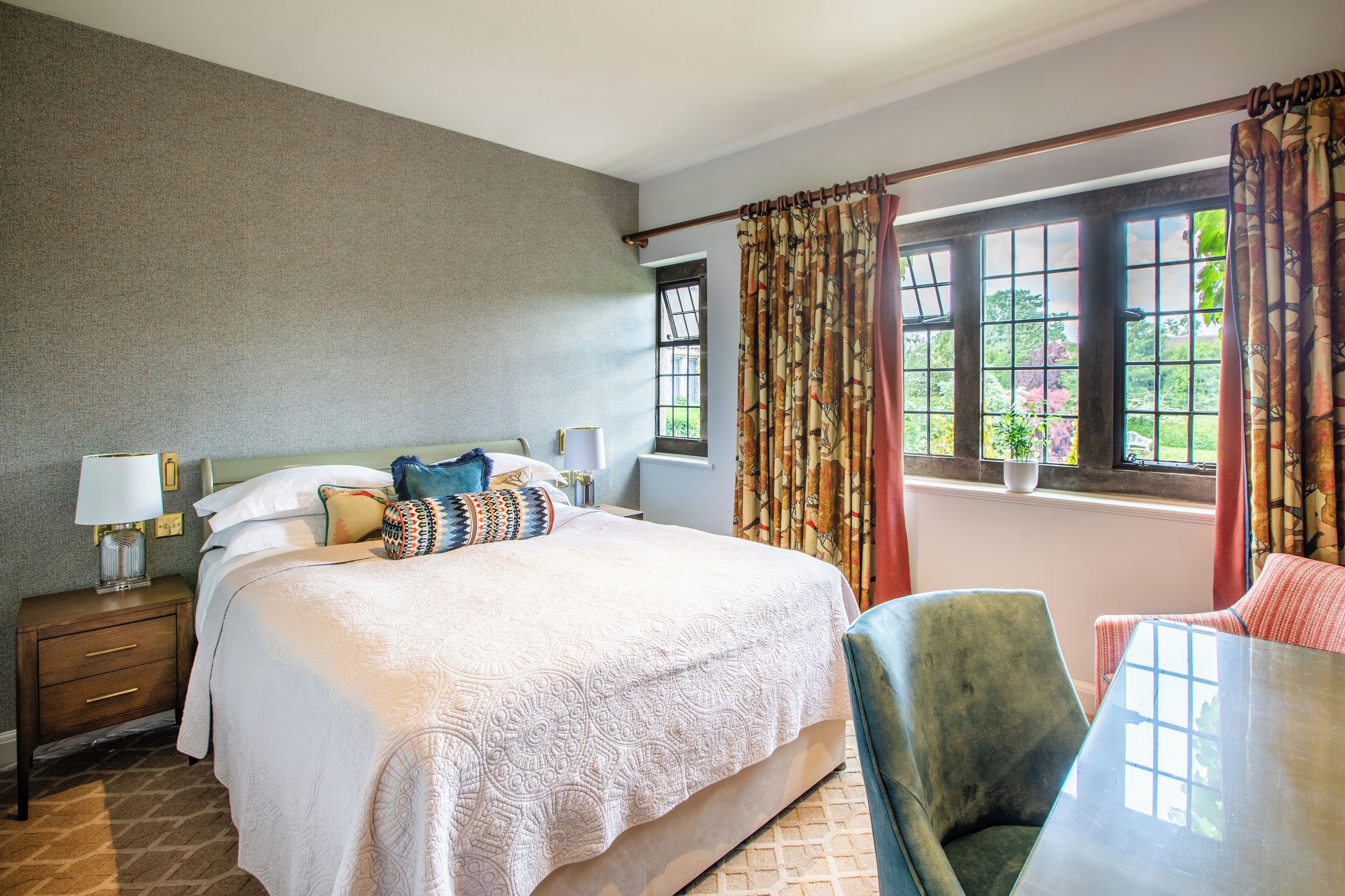 A hotel room with a large bed, a nightstand with a lamp, a chair, a table, and a window with curtains.