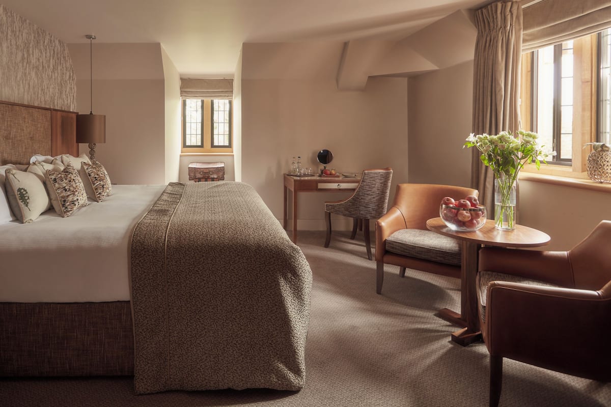 Habitación de hotel con una cama grande, un escritorio con una silla, una mesita con un florero y una ventana con cortinas.