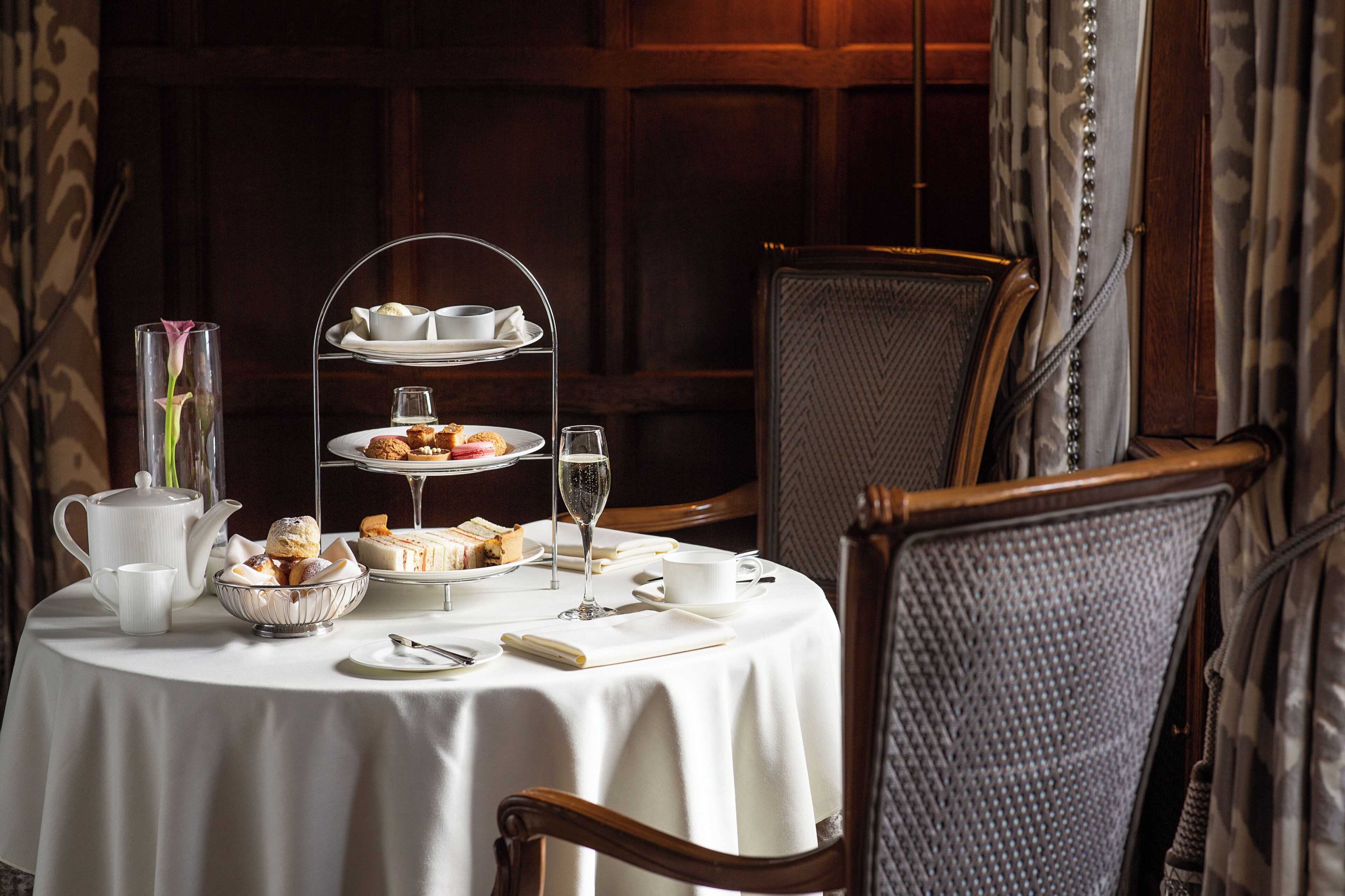 A round table with a white tablecloth, a tiered tea service, a vase with a single flower, a bowl of pastries, a teapot, and a cup.