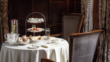 A round table with a white tablecloth, a tiered tea service, a vase with a single flower, a bowl of pastries, a teapot, and a cup.