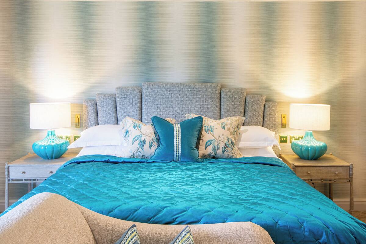 Una cama bien tendida con un edredón azul, dos mesitas de noche con lámparas y un cabecero con textura.